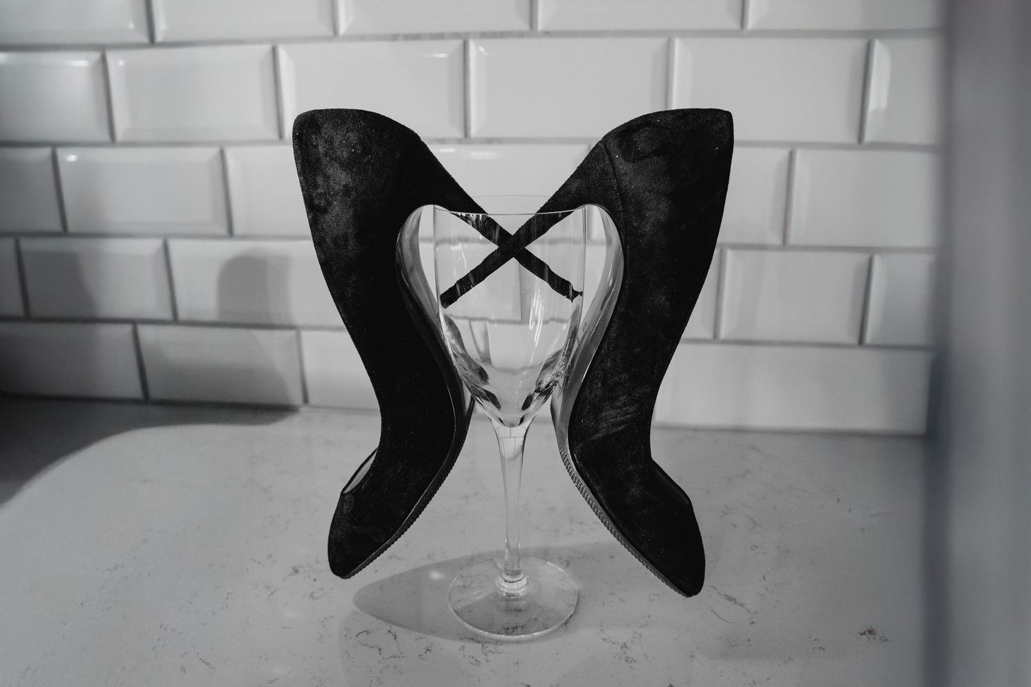 Black and white photo of two black high heel shoes balanced elegantly in a wine glass against white subway tiles.