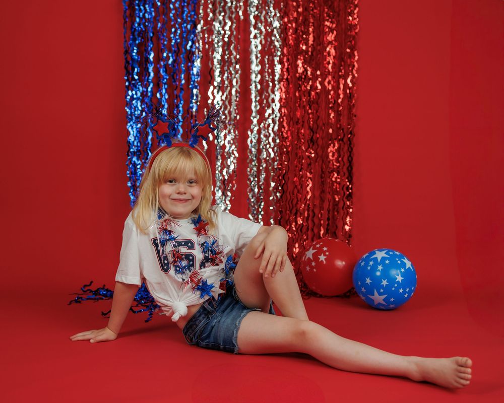 Child poses on red floor against sparkly red, white and blue backdrop with patriotic themed outfit and balloon decoration.