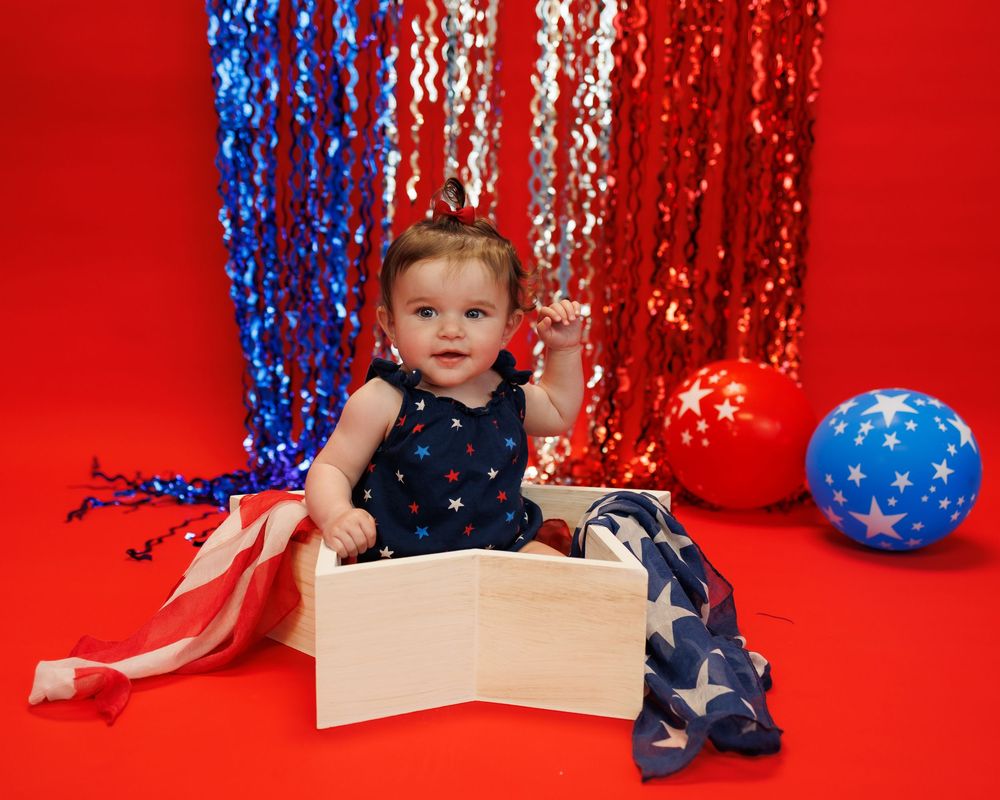 Patriotic themed photoshoot featuring red backdrop, blue and white tinsel decorations, and a white star prop.