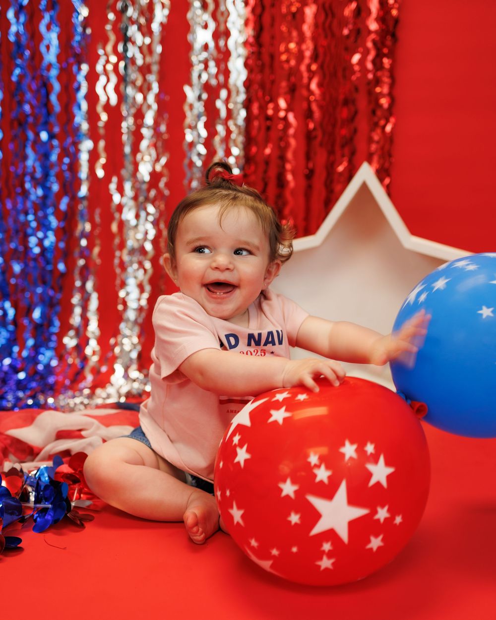 Festive patriotic scene with red and blue sequin backdrop, star balloons, and a smiling baby in a pink outfit on red floor.