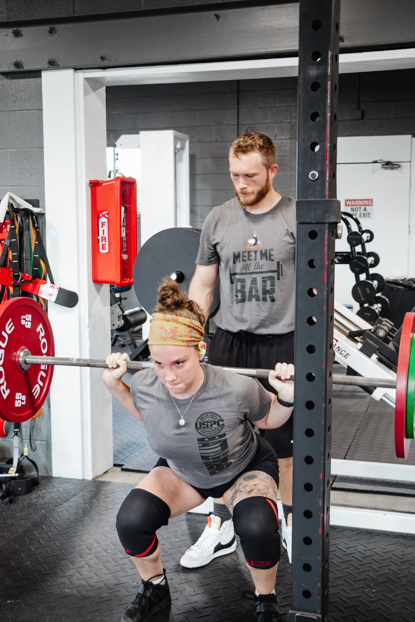 Woman performing a heavy back squat with a barbell as a male spotter stands behind her for support in a gym setting.