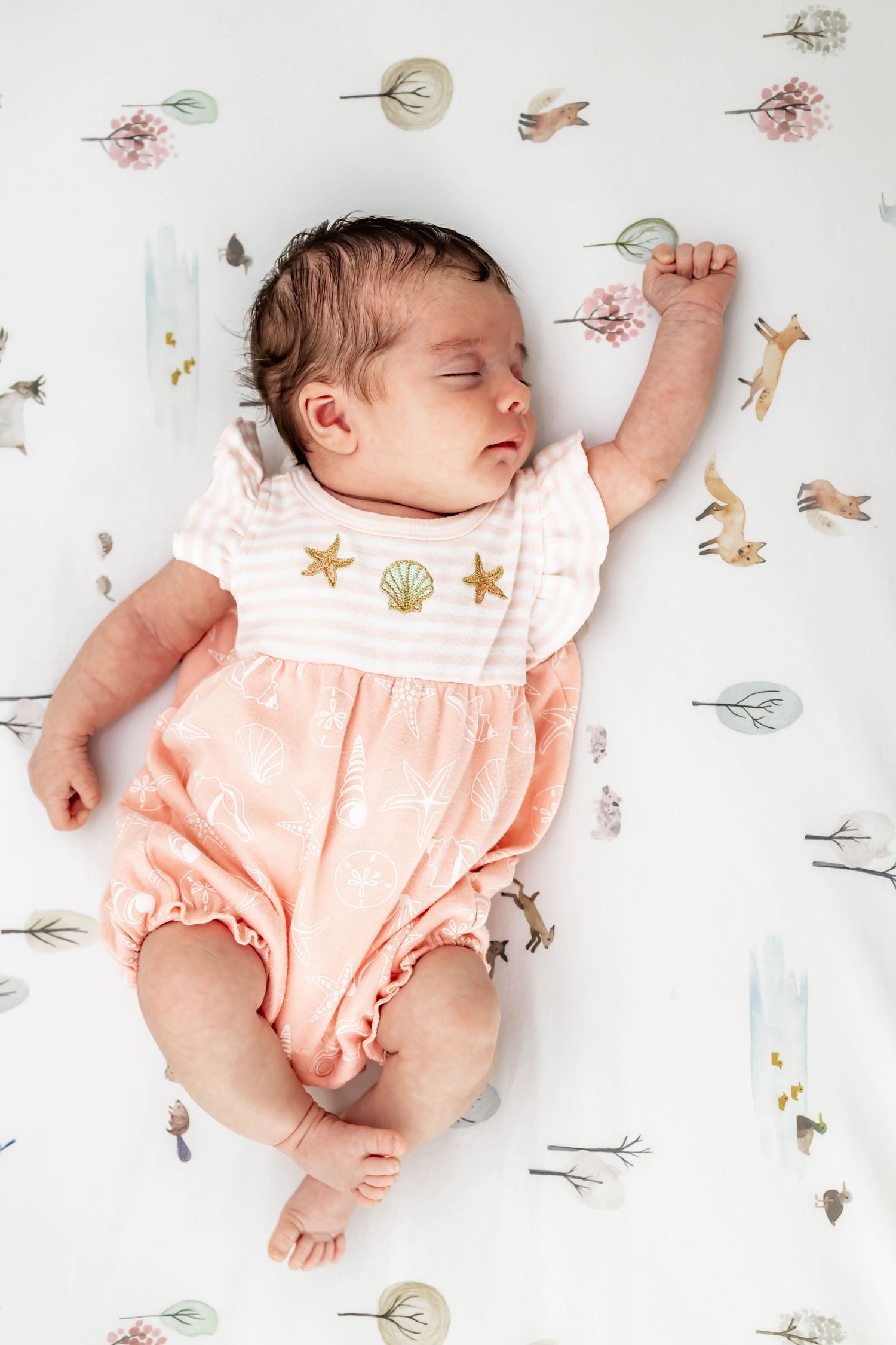 Sleeping baby wearing a peachy pink romper lies on white bedding with delicate floral and nature print pattern.