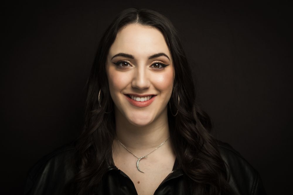 Portrait of a person with dark wavy hair and a warm smile against a dark background wearing a black top.