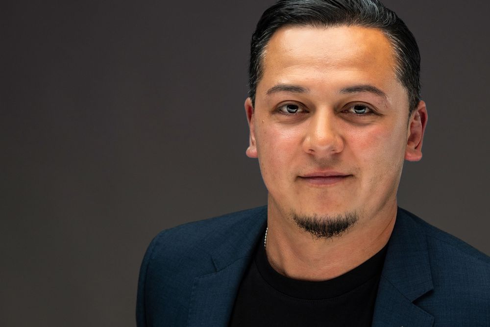 Professional headshot portrait against dark background showing person in navy blazer with facial hair.