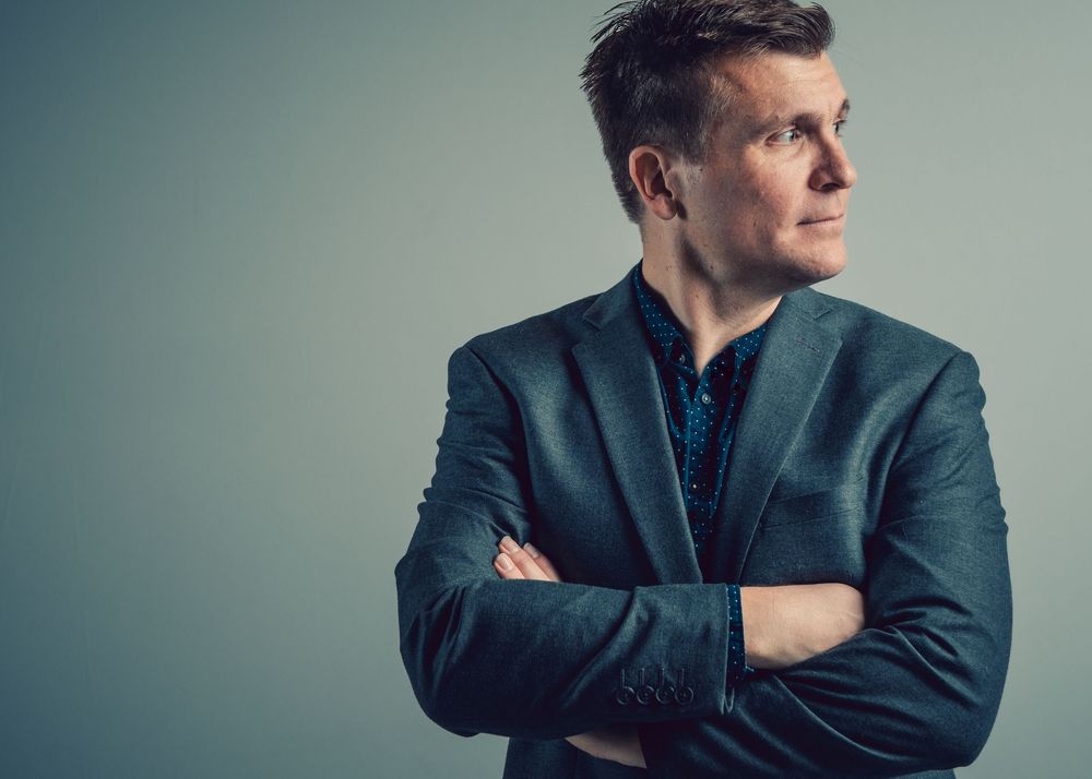Professional portrait of a person in a teal suit jacket standing with arms crossed looking contemplatively to the side.