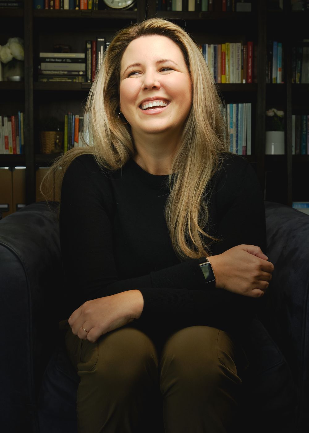 Someone sits smiling warmly on a dark couch with bookshelves visible in the background, wearing a black sweater.