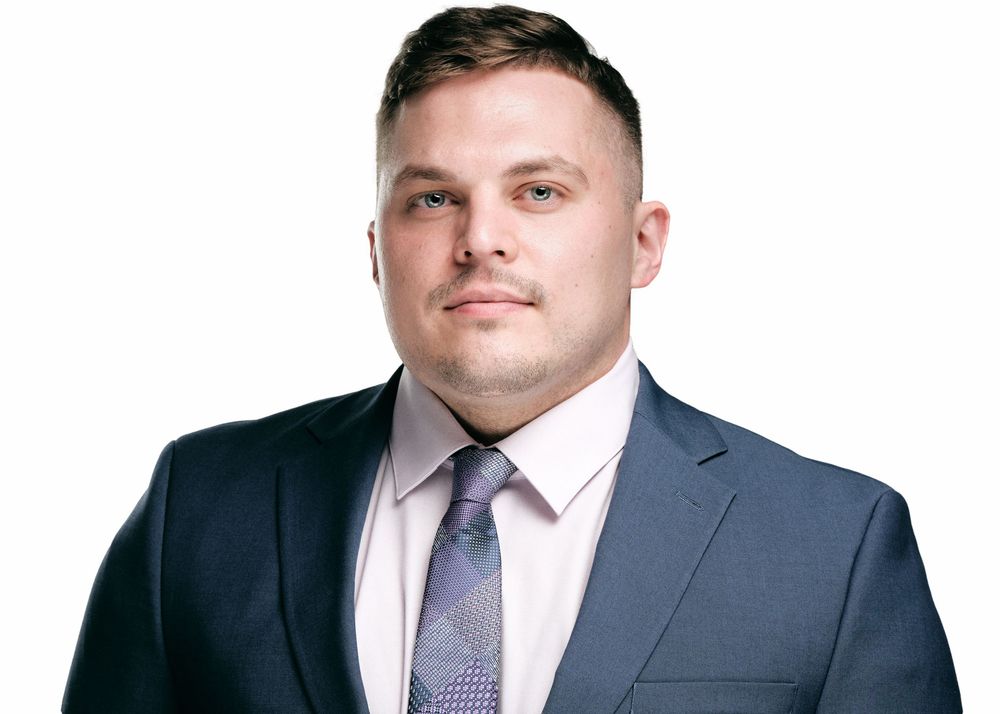 Professional headshot of person in navy suit and purple tie against neutral background.
