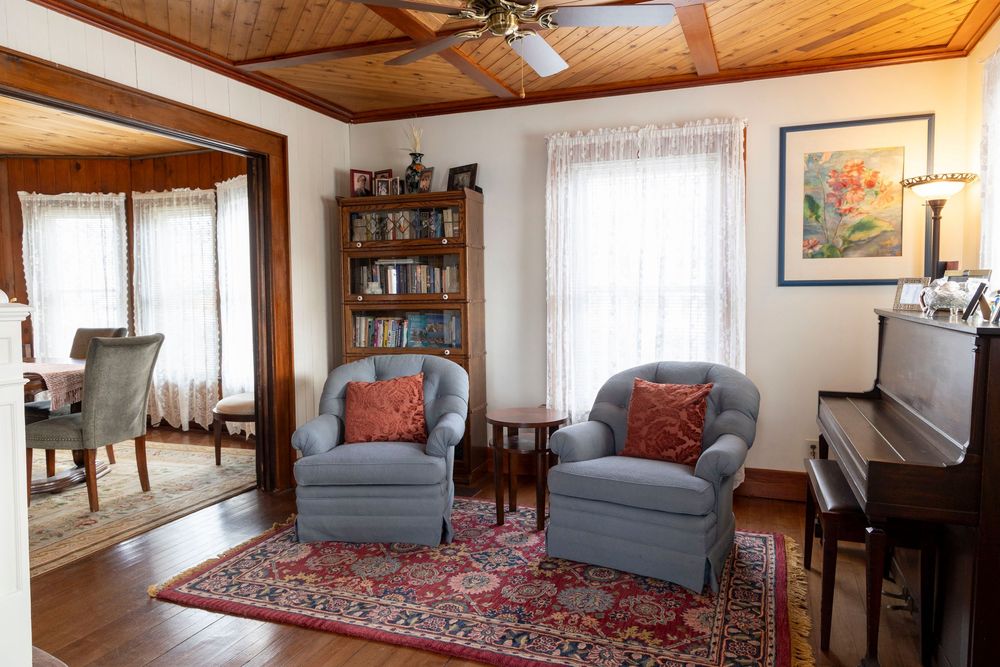 Cozy living room with blue armchairs, oriental rug, and upright piano under wooden ceiling beams.