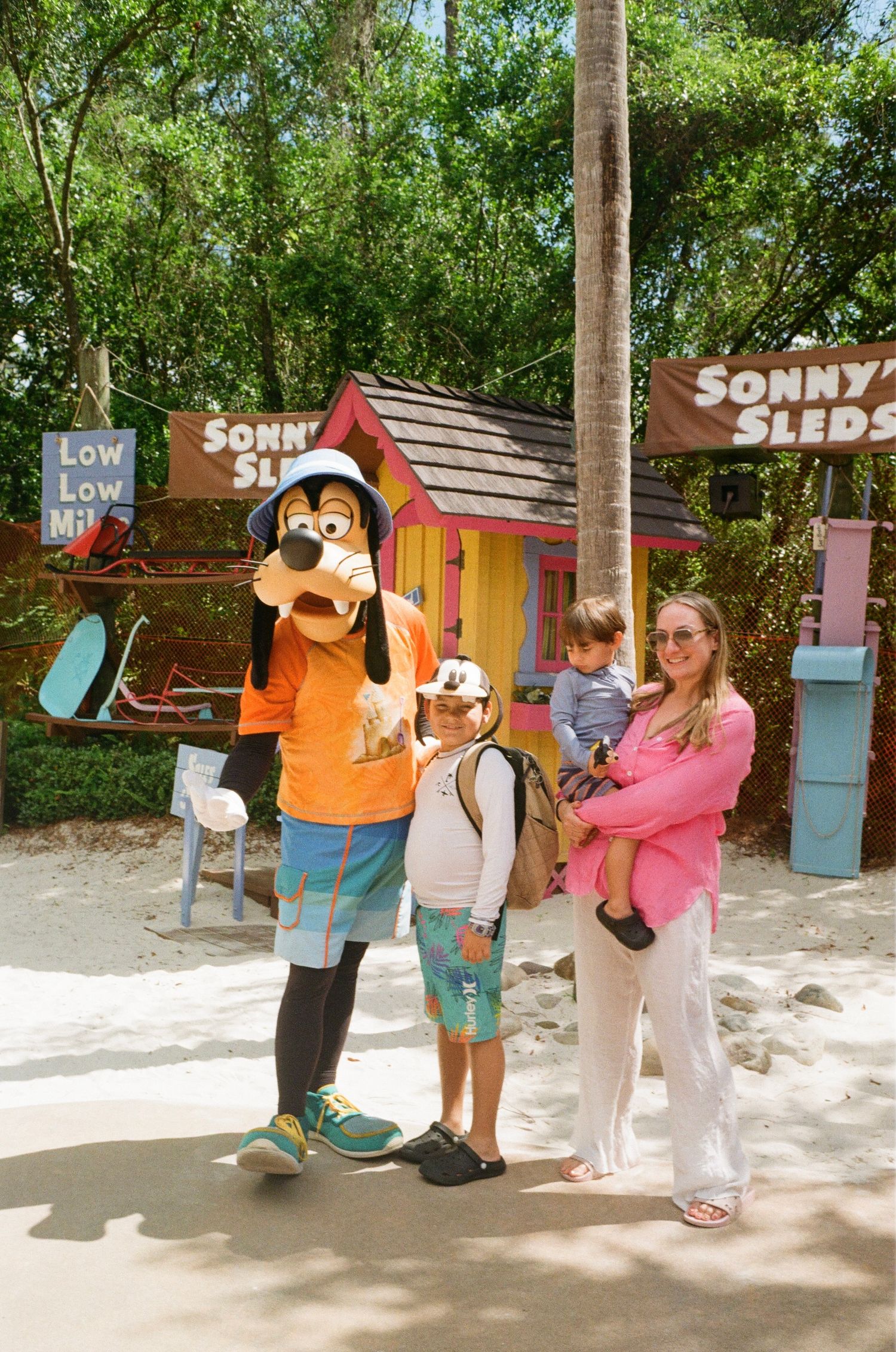 Family poses with Goofy character at Disney theme park with small wooden structures in the background.