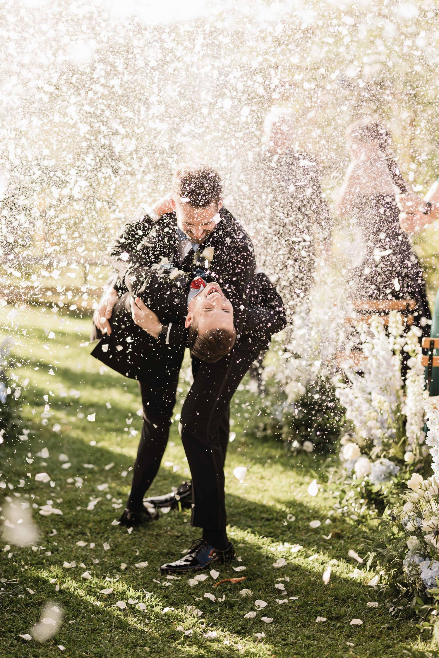 A couple shares a romantic moment surrounded by swirling flower petals in a sun-drenched garden setting.