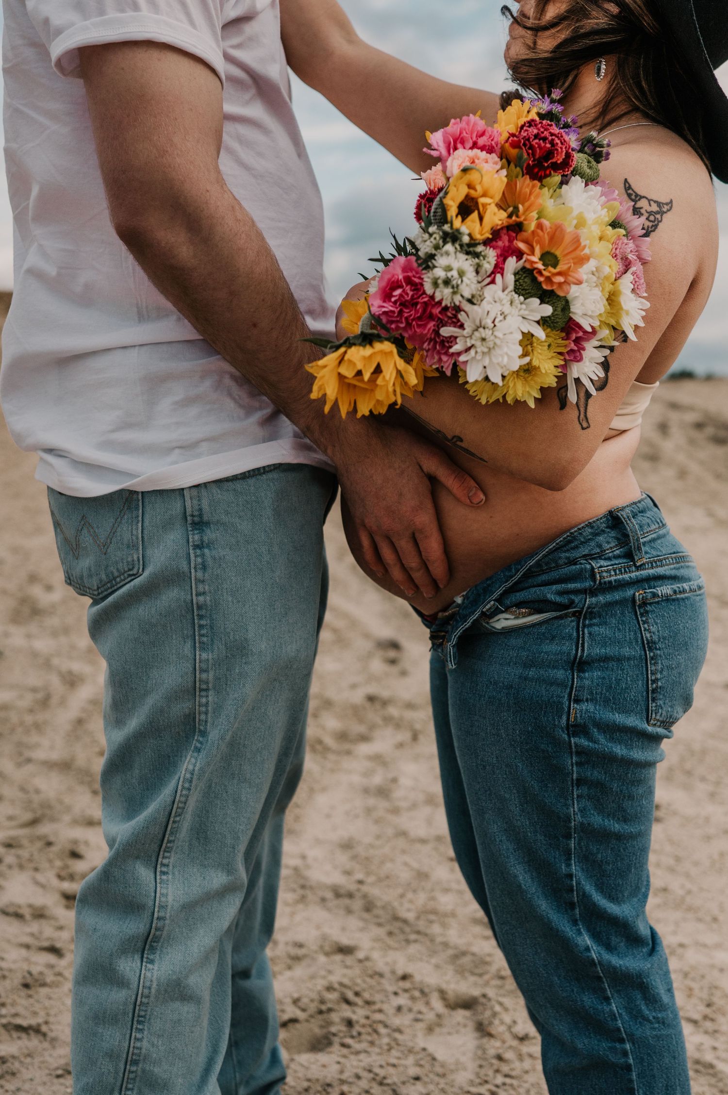 A colorful floral bouquet with sunflowers and roses is held against a woman's pregnant belly as two people stand close on sandy ground.