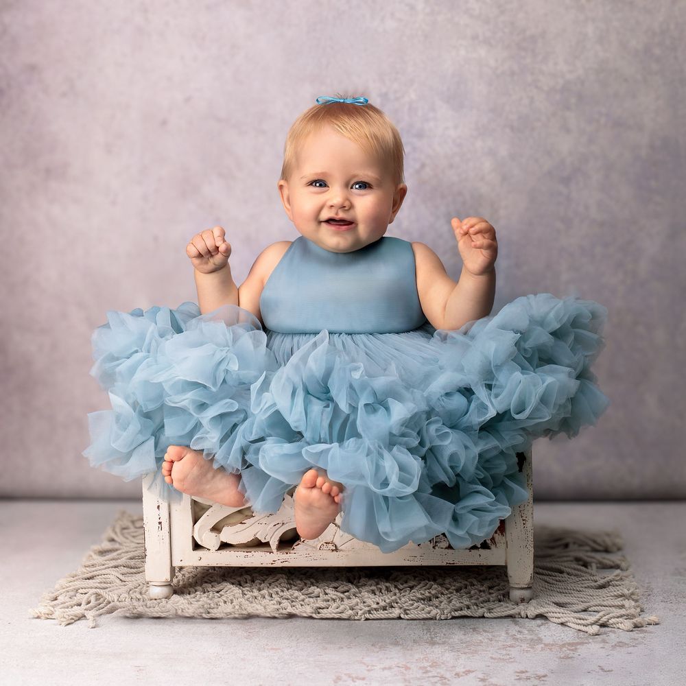 Baby Girl sits on a white wooded bed prop wearing a couture blue dress smiling, by Sydney family studio photographer Caz Captures.