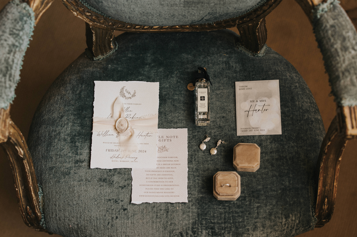 Wedding stationery suite with invitations, ring box and perfume bottle laid out on dark velvet chair.