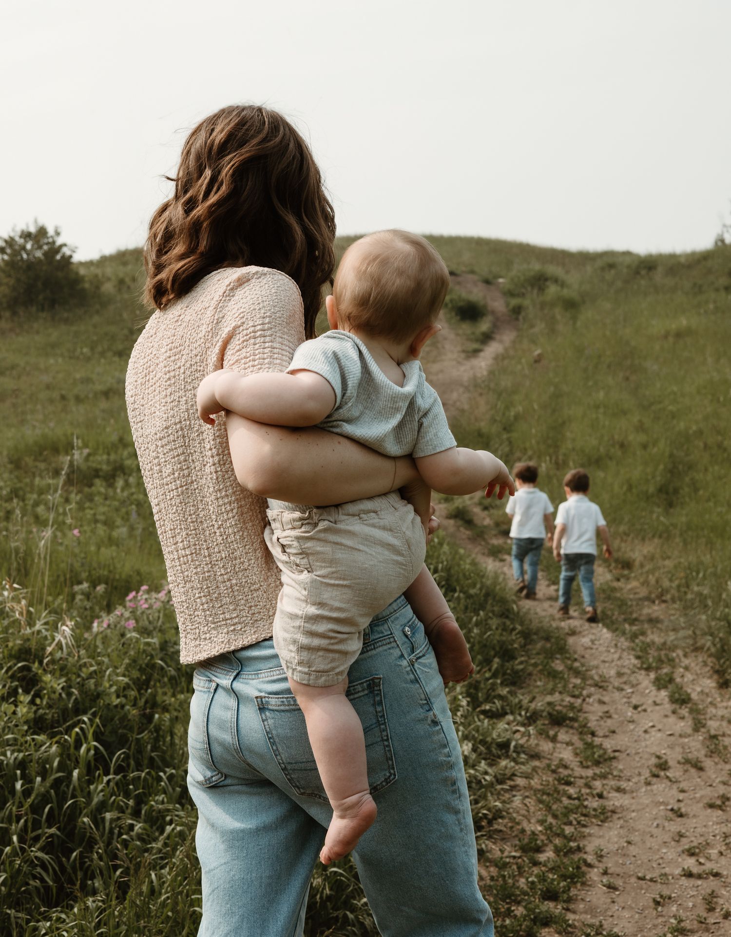 Mother holding baby while watch her boys run down a path between hills in Nose Hill Park in Calgary