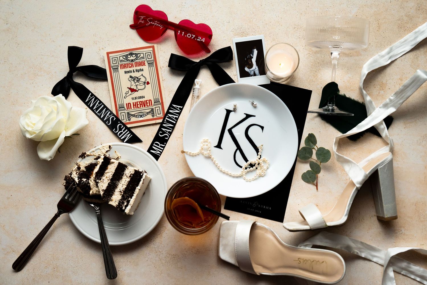 Elegant wedding flatlay with cake slice, monogram plate, shoes, tickets, and red bow against neutral backdrop.