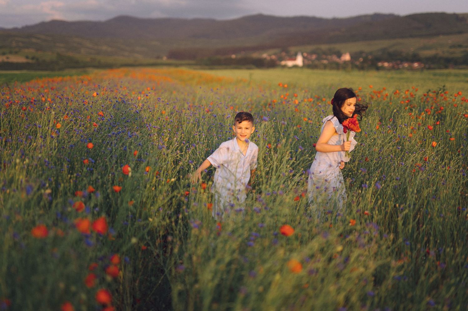 Two people running playfully through a tall grass meadow filled with vibrant red poppies at sunset.