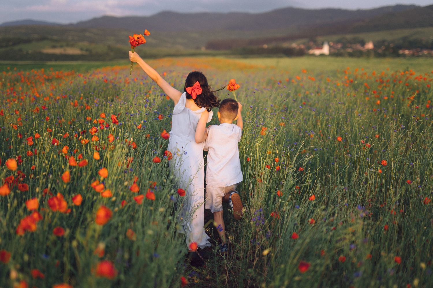 Two figures stand amid a vibrant field of red poppies at sunset with mountains in the distance.
