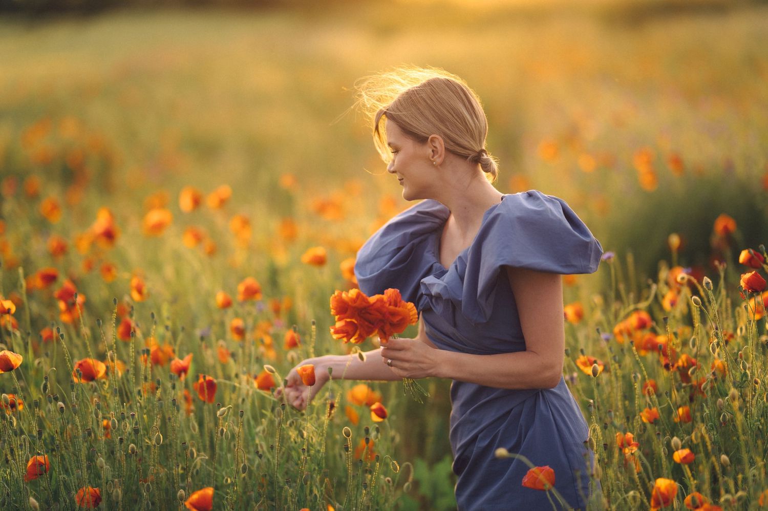 Side view of a person in a flowing blue dress walking through a vibrant field of orange poppies at sunset.