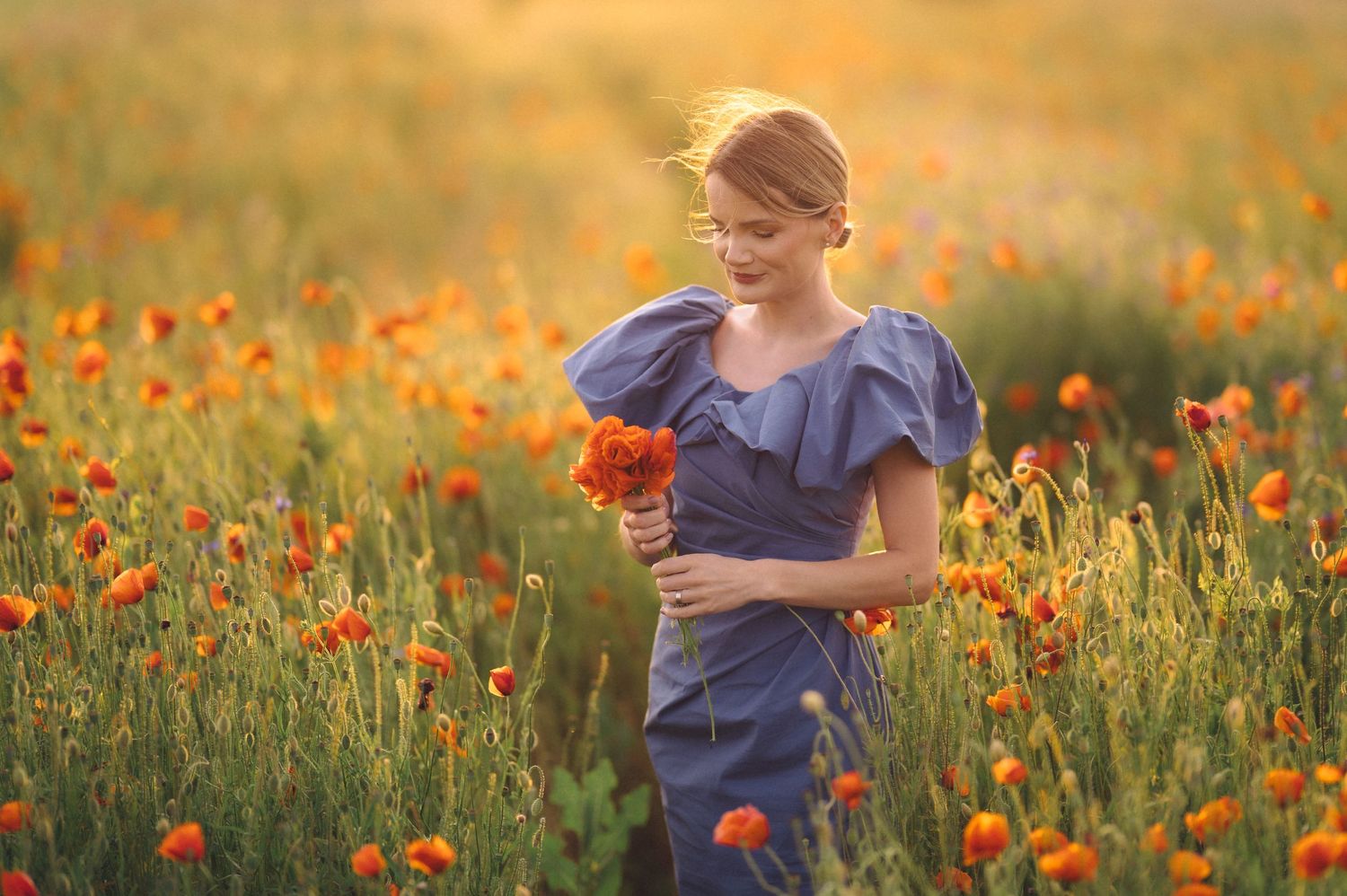 A person in a blue dress stands in a vibrant field of orange poppies at golden hour.