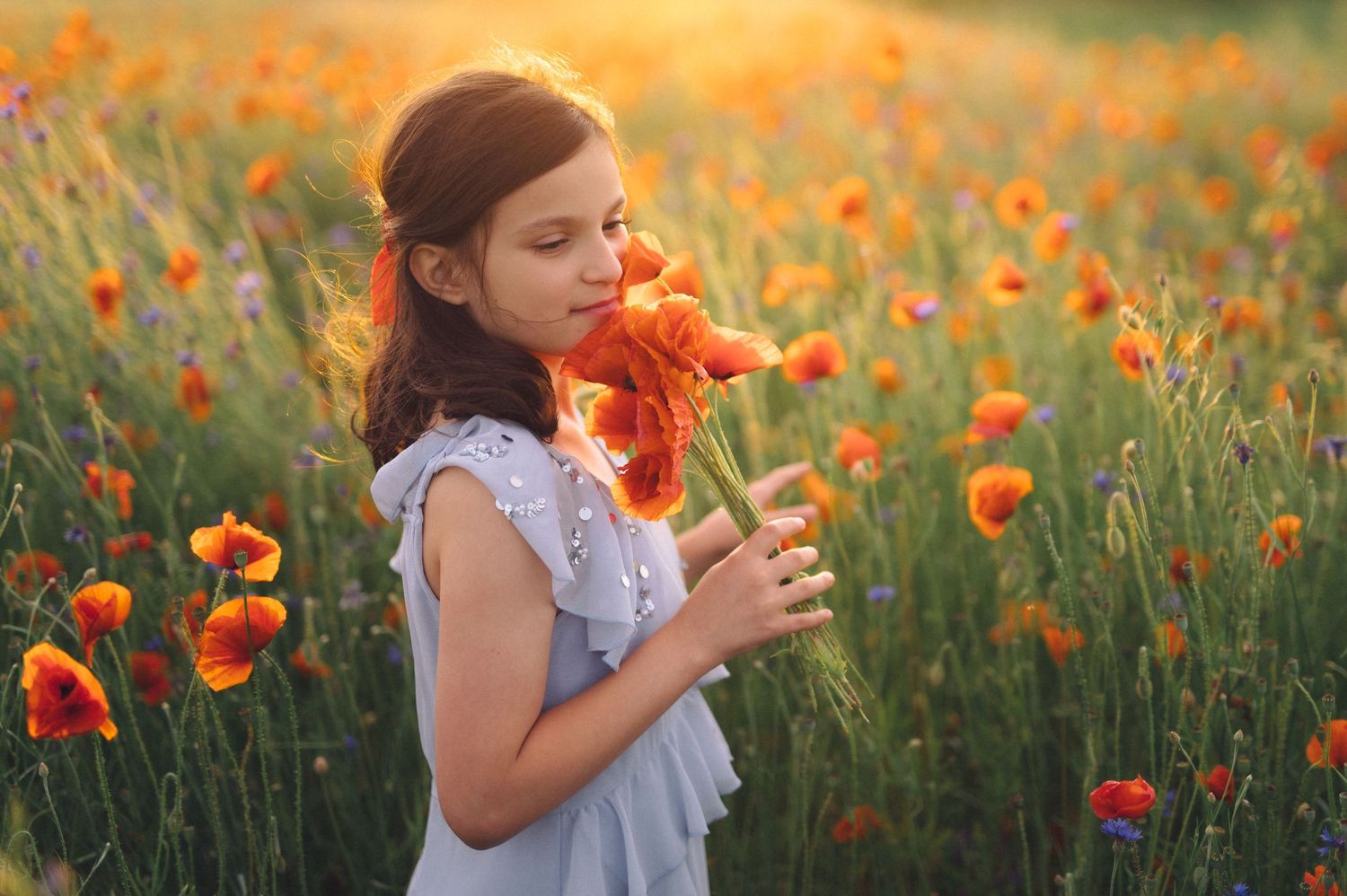 A person in a white dress smells vibrant orange poppies in a dreamy summer meadow at golden hour.