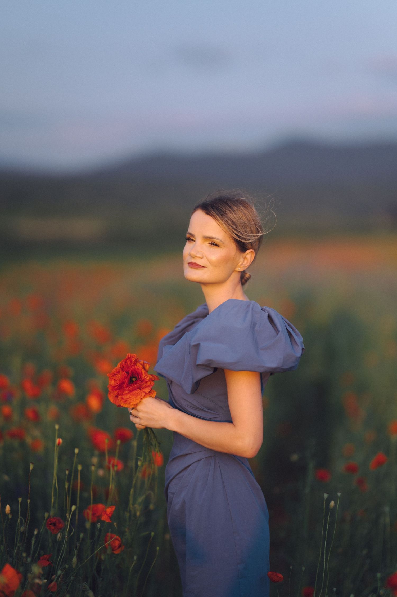 A romantic scene in a poppy field at sunset showing a figure in a blue dress with puff sleeves holding red flowers.