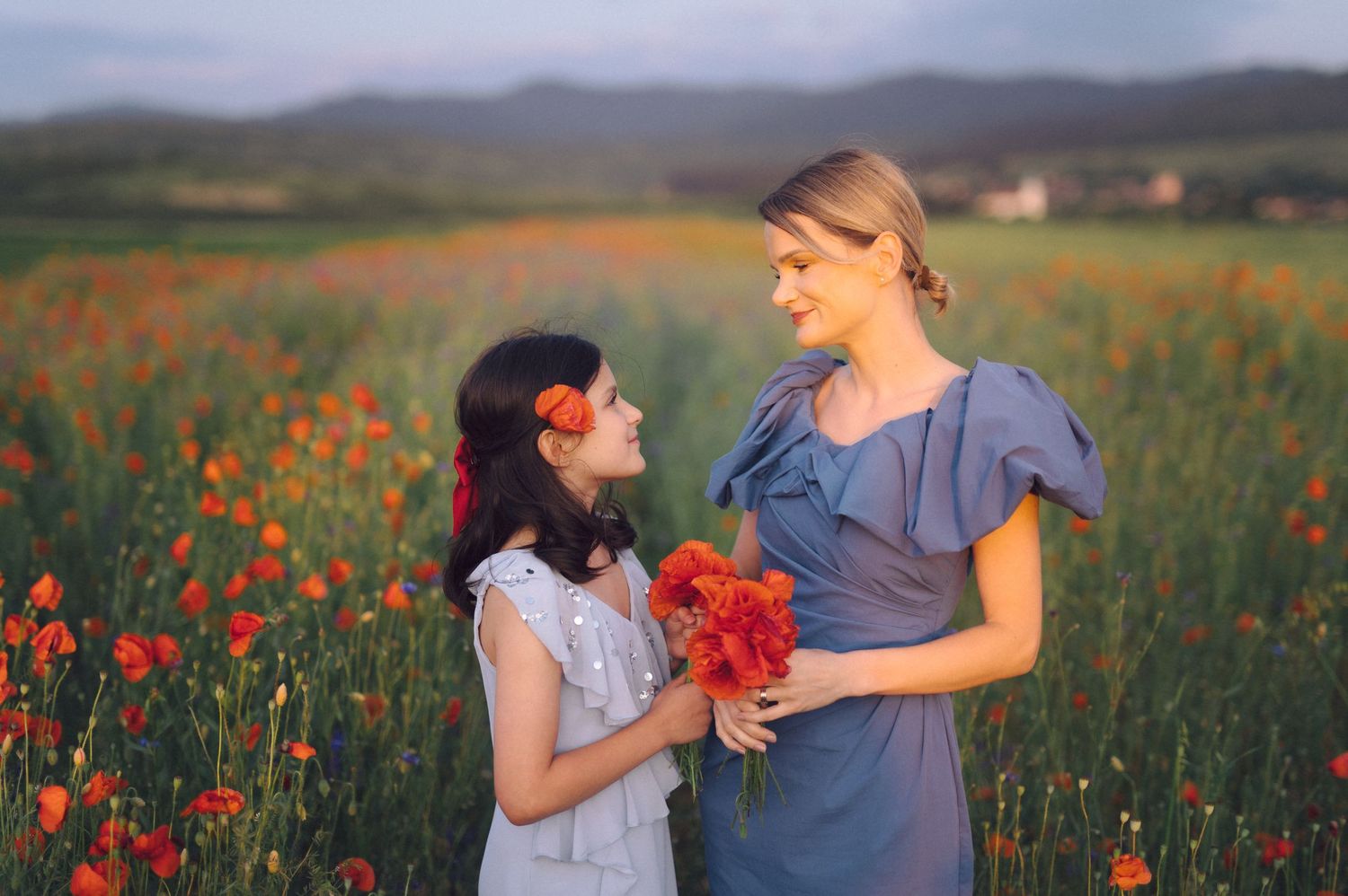 Two people stand together in a vibrant field of red poppies during golden hour in the countryside.