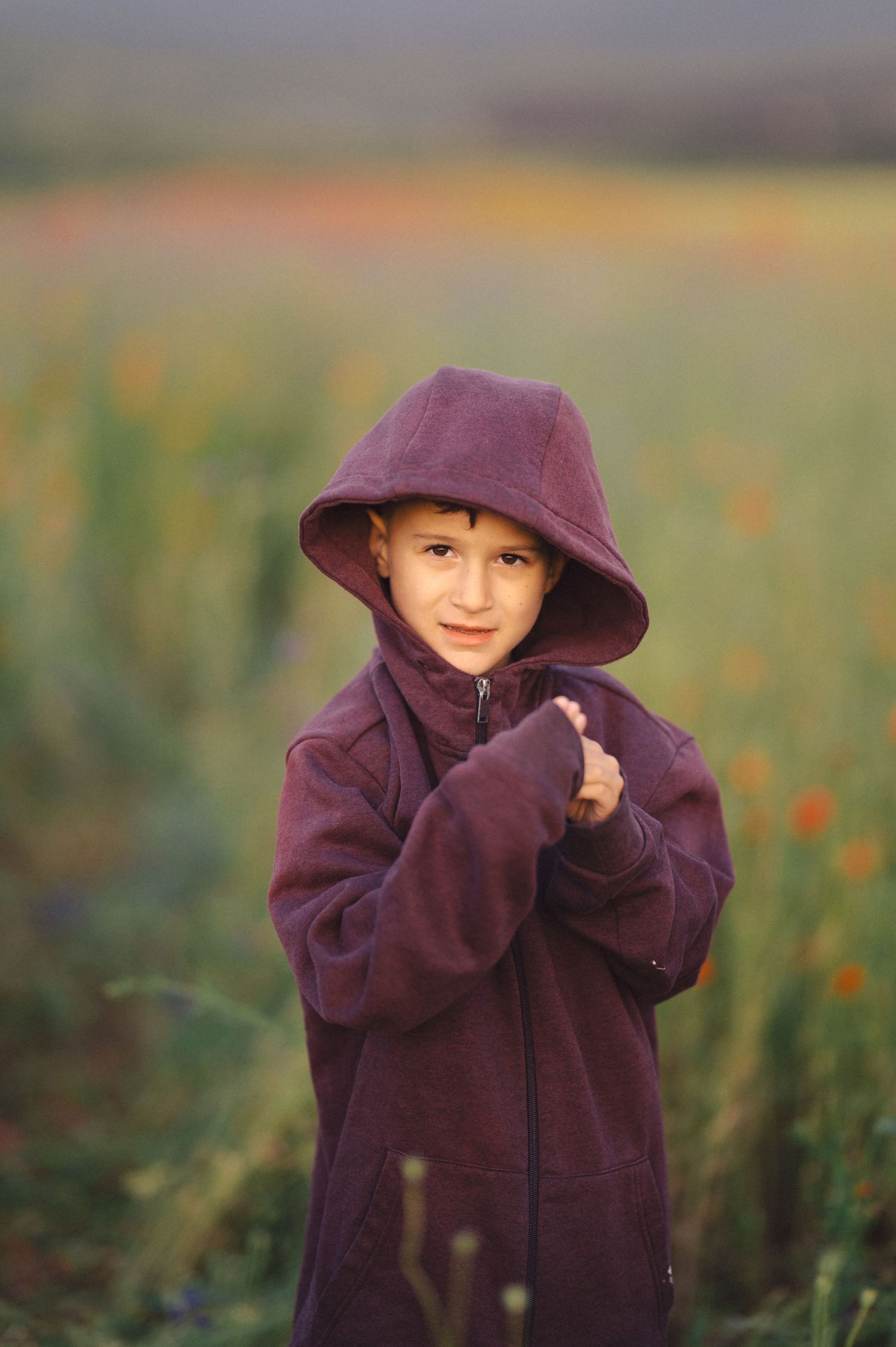 Child in orchid hoodie plays peekaboo in blooming meadow as golden sunlight illuminates the background.