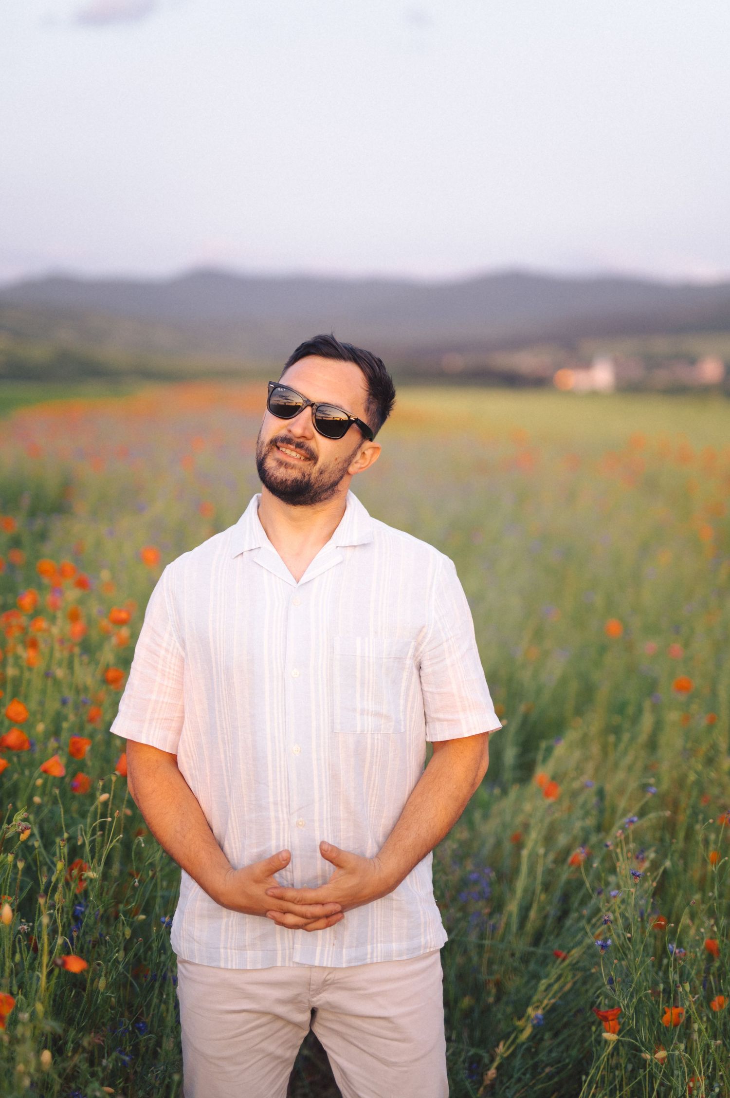 Person in white outfit stands in vibrant field of red poppies with rolling hills in the background during golden hour.