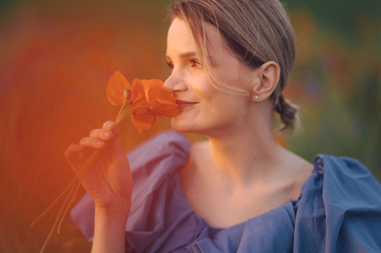 A person in a blue dress smelling an orange flower with a soft dreamy sunset glow in the background.