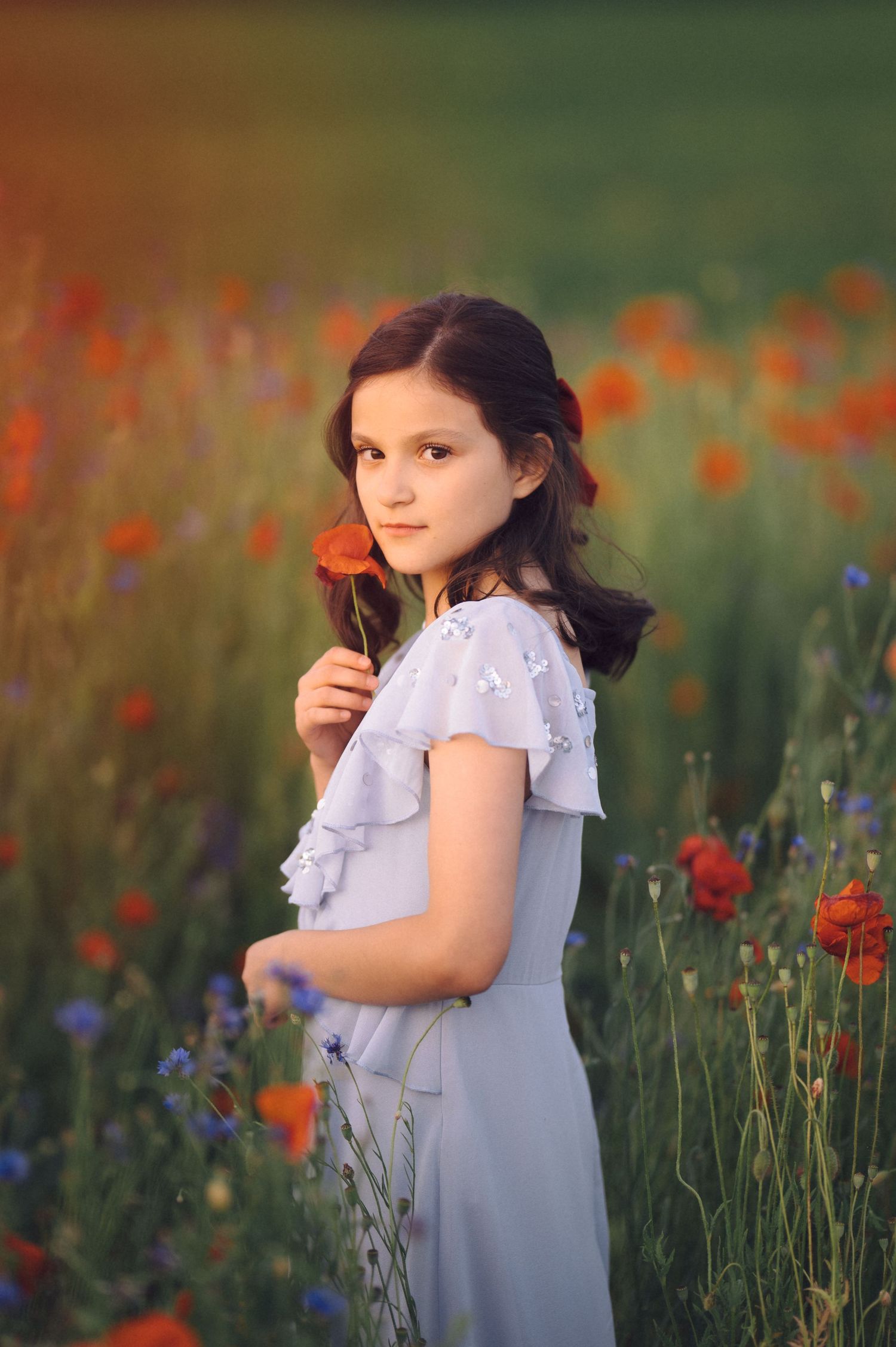 A person in a light blue ruffled dress stands in a field of vibrant red poppies and wildflowers at golden hour.