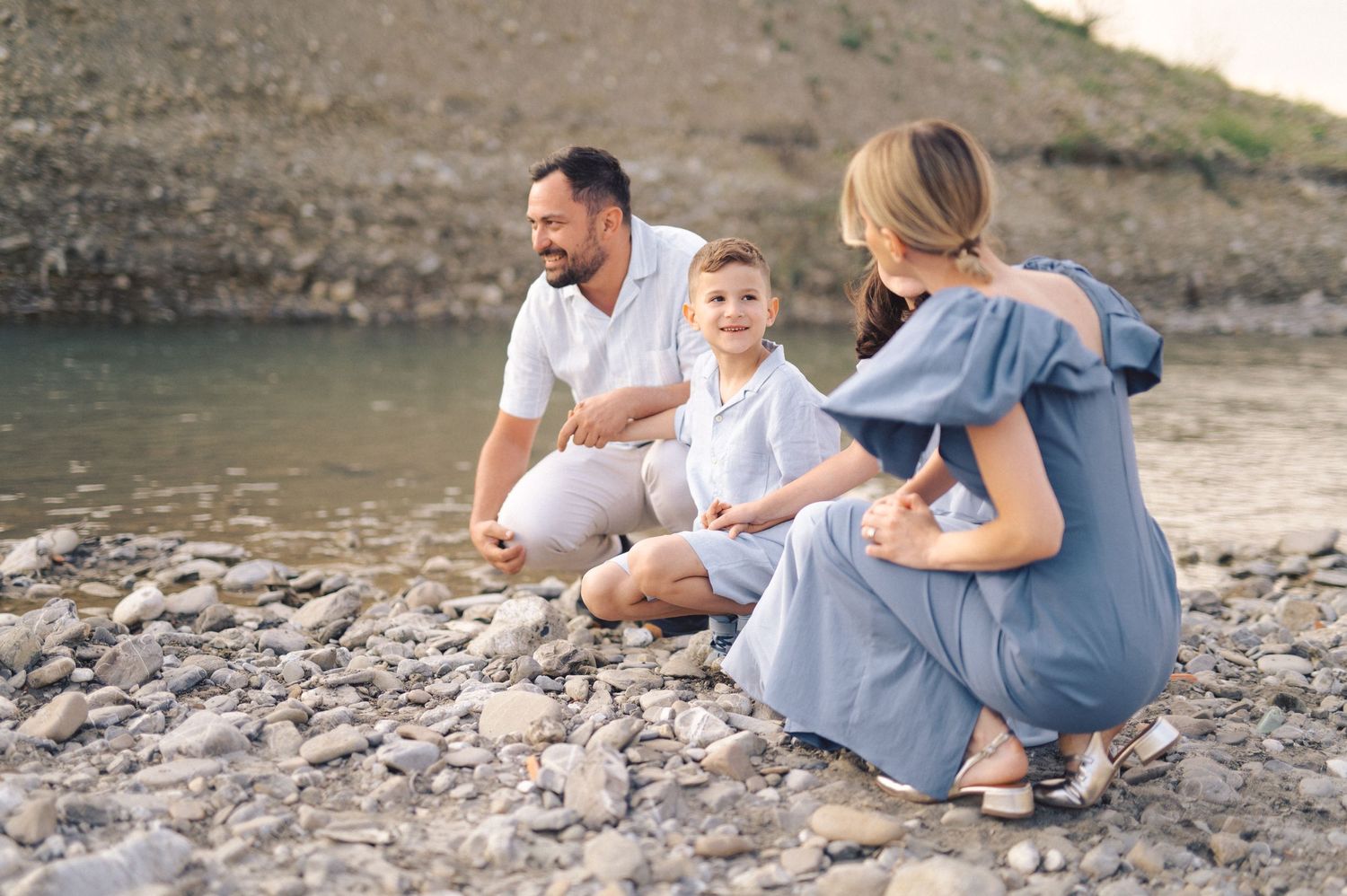 A family in coordinated blue outfits sits on a rocky shoreline near the water on a sunny afternoon.