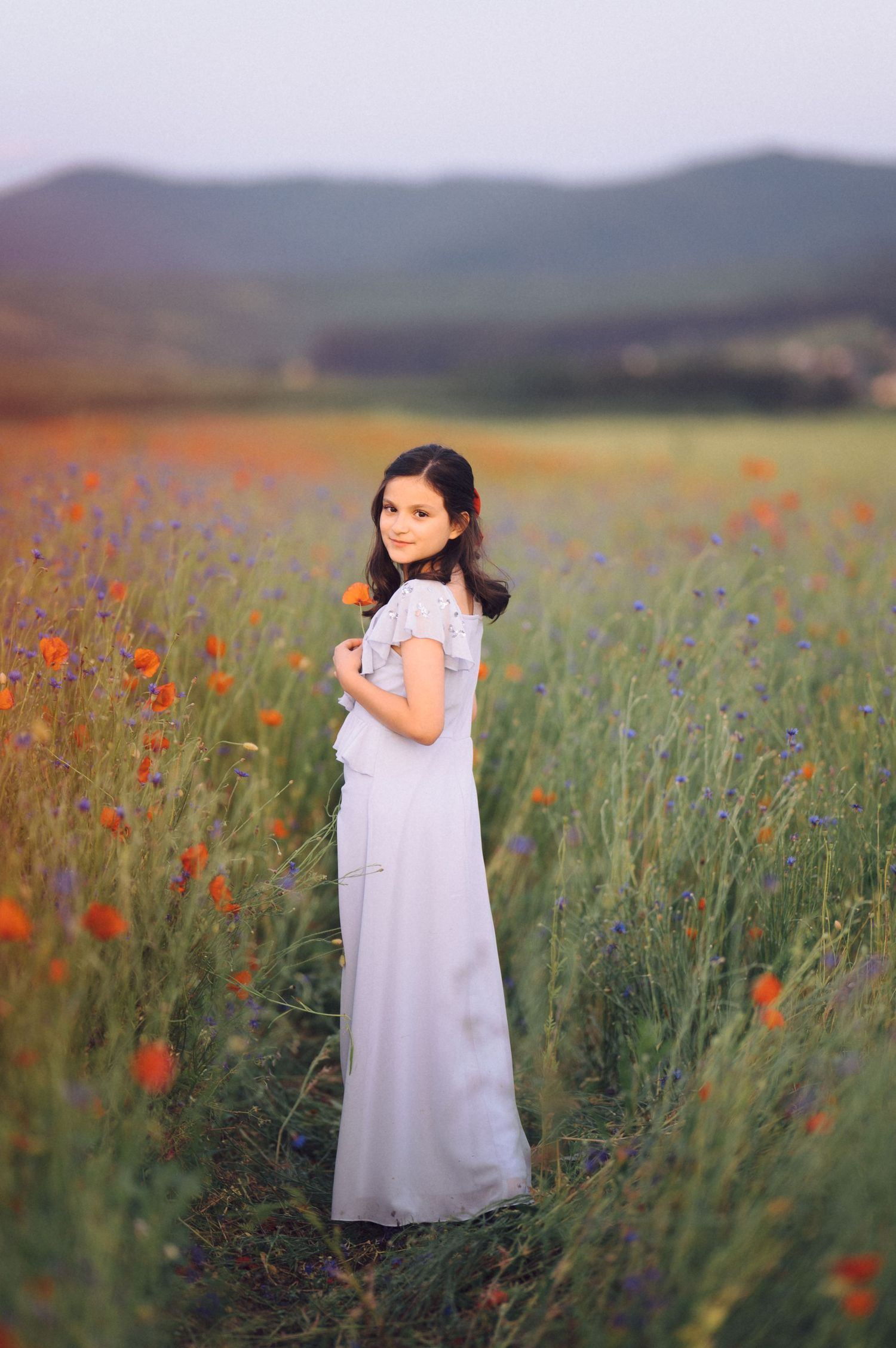 A person in a flowing white dress stands in a serene meadow with red poppies and tall grass against mountain scenery.