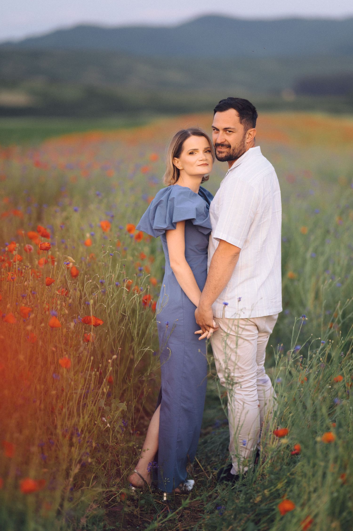 A couple embraces in a field of red poppies against a mountain backdrop during golden hour.