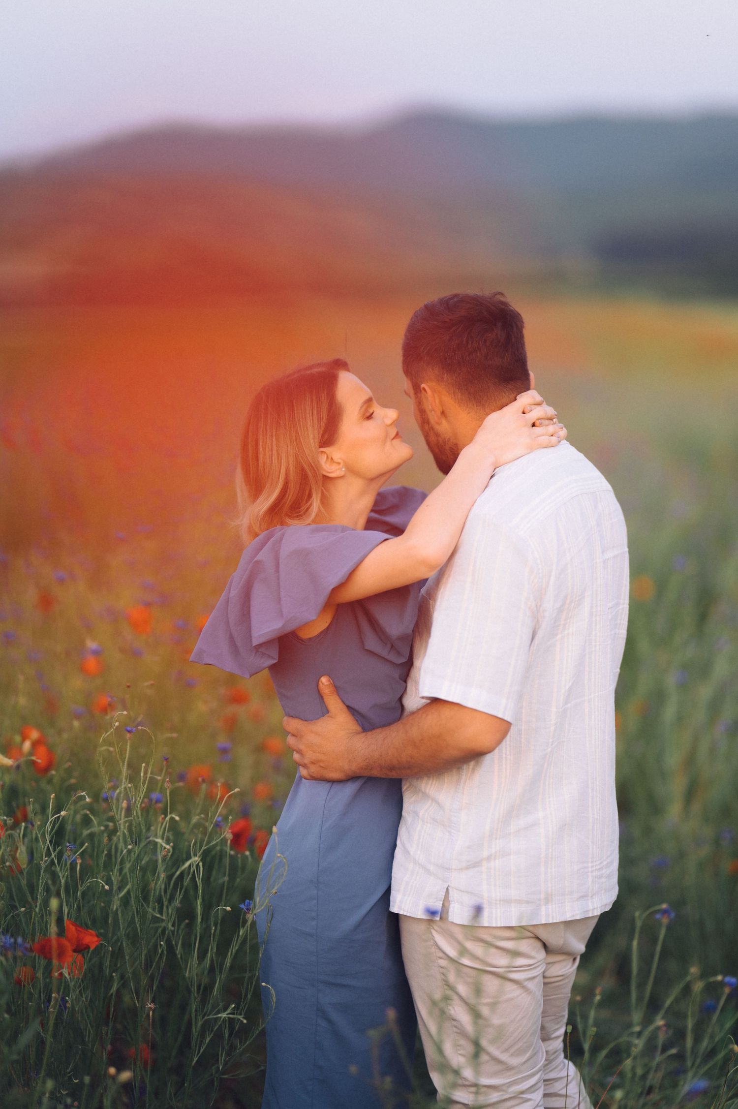 Romantic couple embracing in a dreamy poppy field at sunset with warm golden light glowing in background.