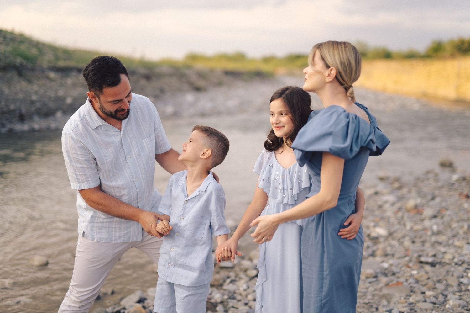 A family in light colored clothing shares a tender moment together while standing on a rocky beach at sunset.