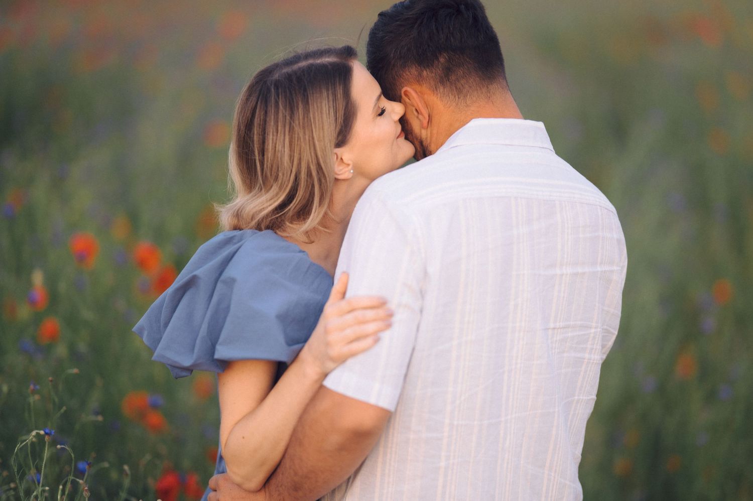 Two people share a tender embrace while standing in a meadow with red poppies during golden hour.