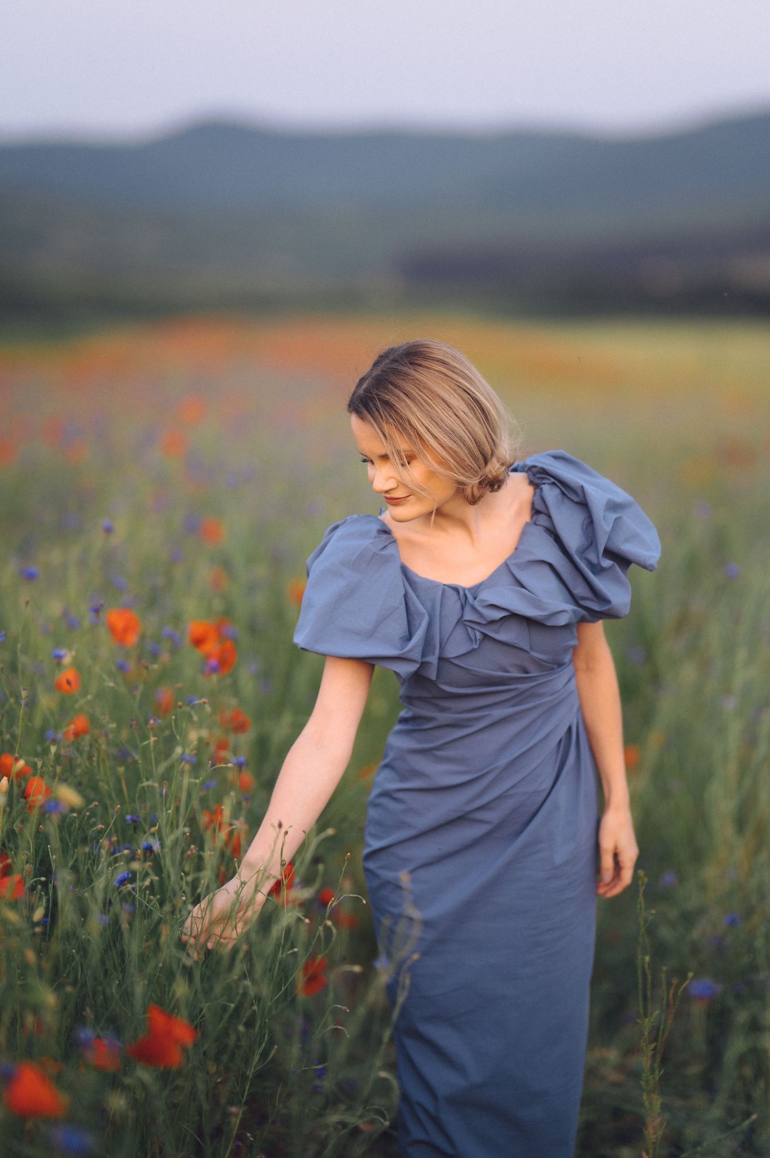 Someone in a blue dress stands gracefully among vibrant red poppies in a serene meadow during golden hour.