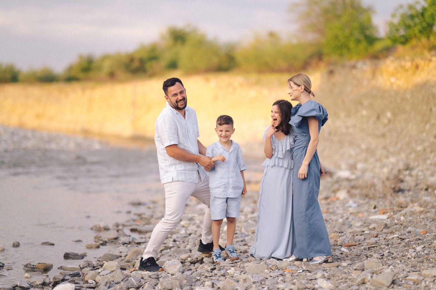 Family relaxes together on a rocky beach during sunset, wearing coordinated blue and white outfits.