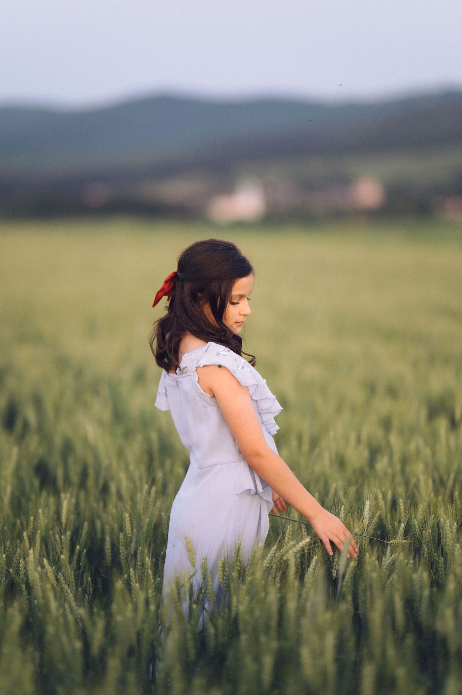 Person in a white dress gently running fingers through tall green wheat stalks in a serene countryside field at sunset.