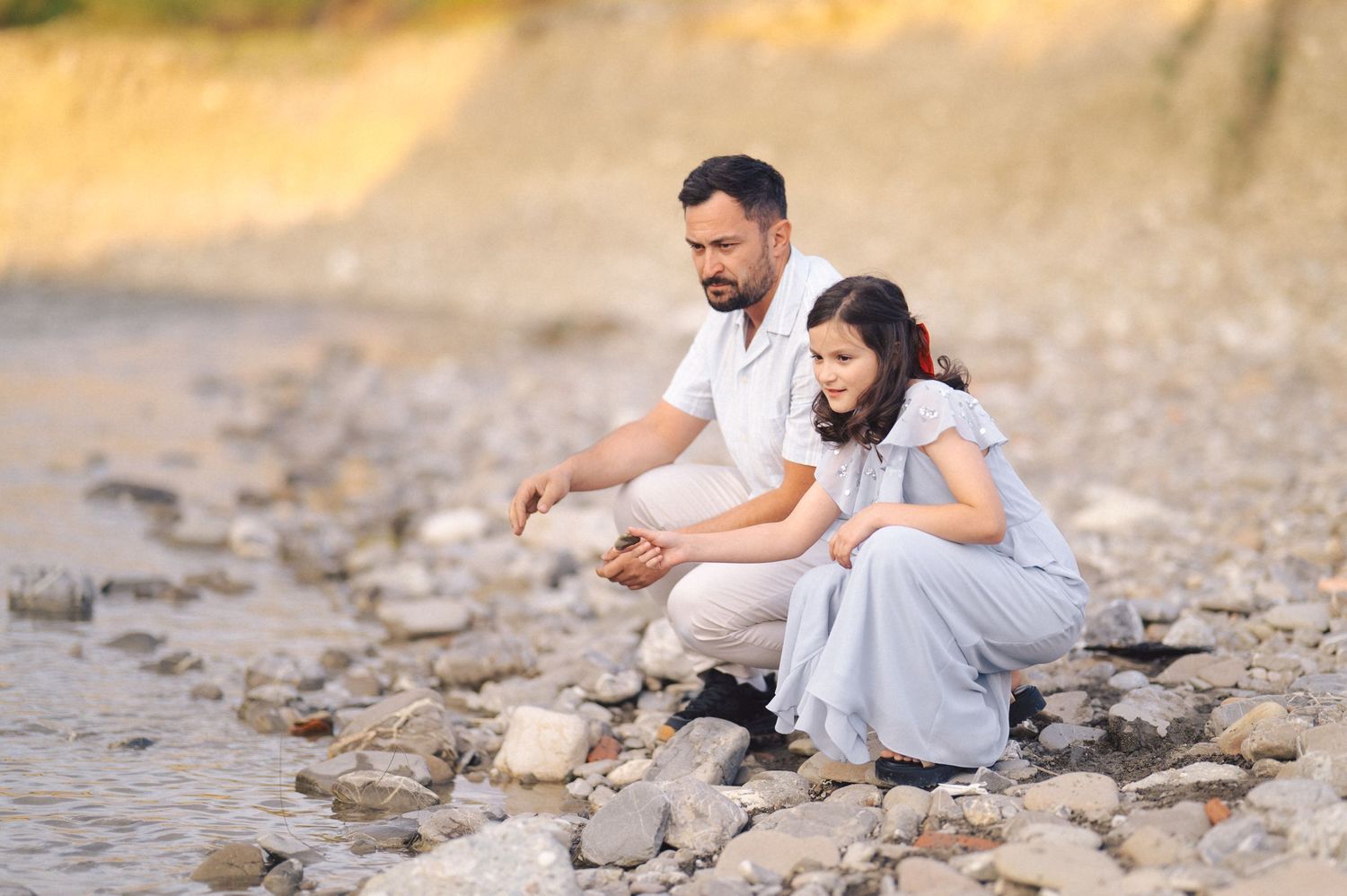 A couple in white outfits sits on a rocky beach shore during a sunset photoshoot.