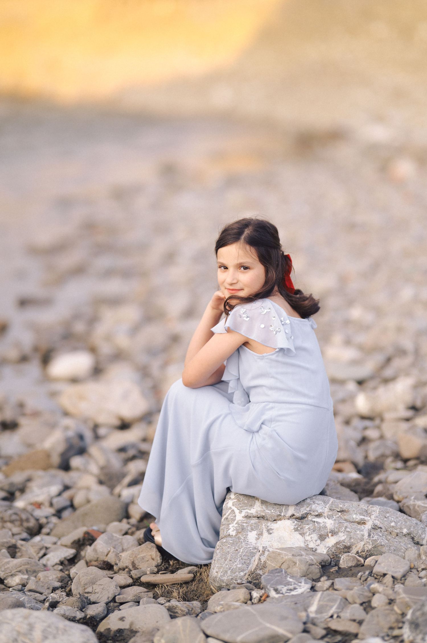 A figure in a light gray outfit sits contemplated on rocky terrain during golden hour.