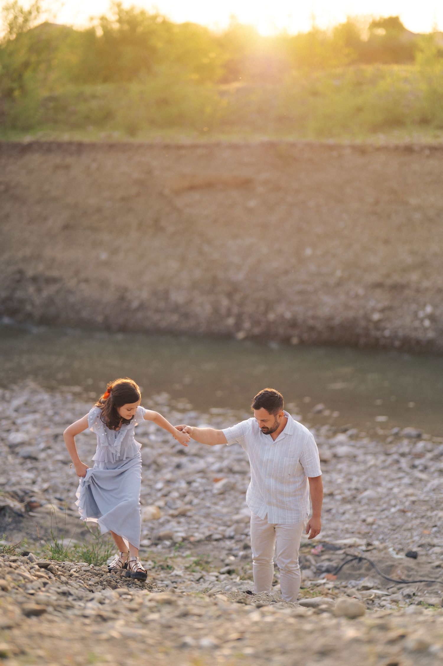 Couple in white outfits walking up rocky riverbank during golden sunset hour.