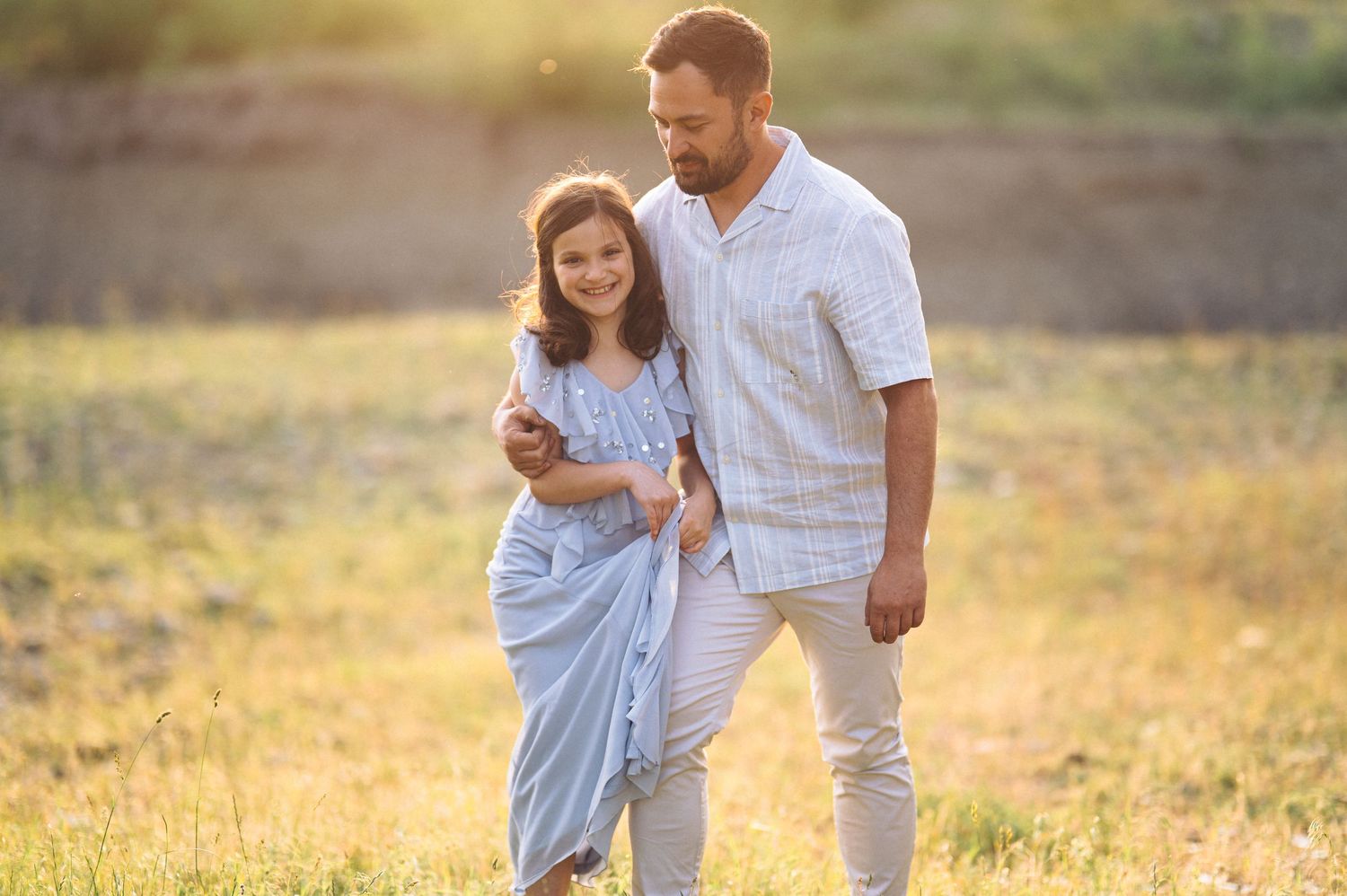Two people in white outfits walk together through a sunlit meadow at golden hour.
