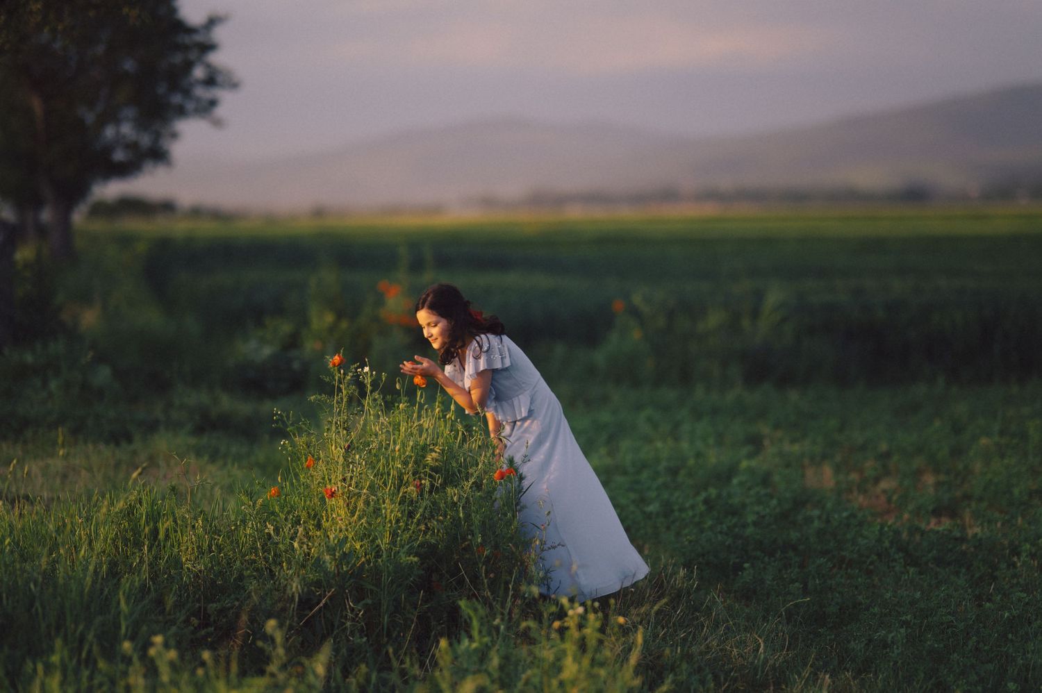 A person in a white dress stands gracefully in a lush green meadow at golden hour with mountains in the distance.