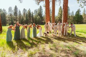 Wedding party in green and neutral dresses poses together in a sunlit forest clearing.