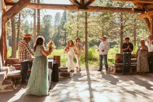 Guests dance and mingle at a rustic outdoor pavilion reception surrounded by pine trees on a sunny afternoon.