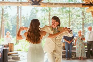 Wedding guests dancing in pairs beneath a wooden pavilion with natural lighting streaming through windows.