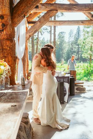 Loving couple embraces under rustic timber beams at an outdoor mountain venue with white curtains and forest views.