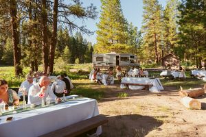 Outdoor wedding reception setup with buffet tables in a sunlit forest clearing surrounded by tall pine trees.