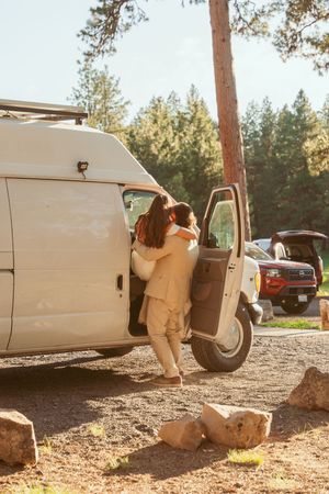Person in beige clothing enters a white van parked in a wooded area during golden hour sunset lighting.
